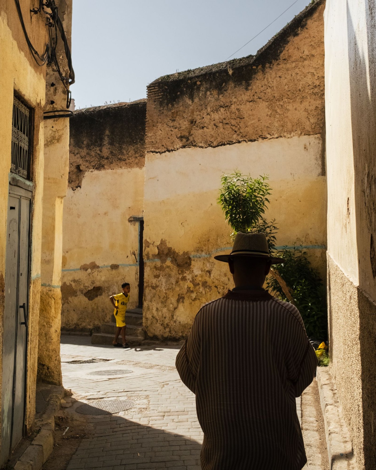 A candid scene of a man in a hat walking through a rustic alleyway with a child playing nearby.