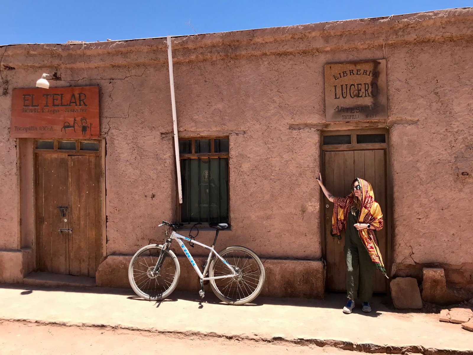A person stands beside a rustic building with a bicycle under the bright sun.