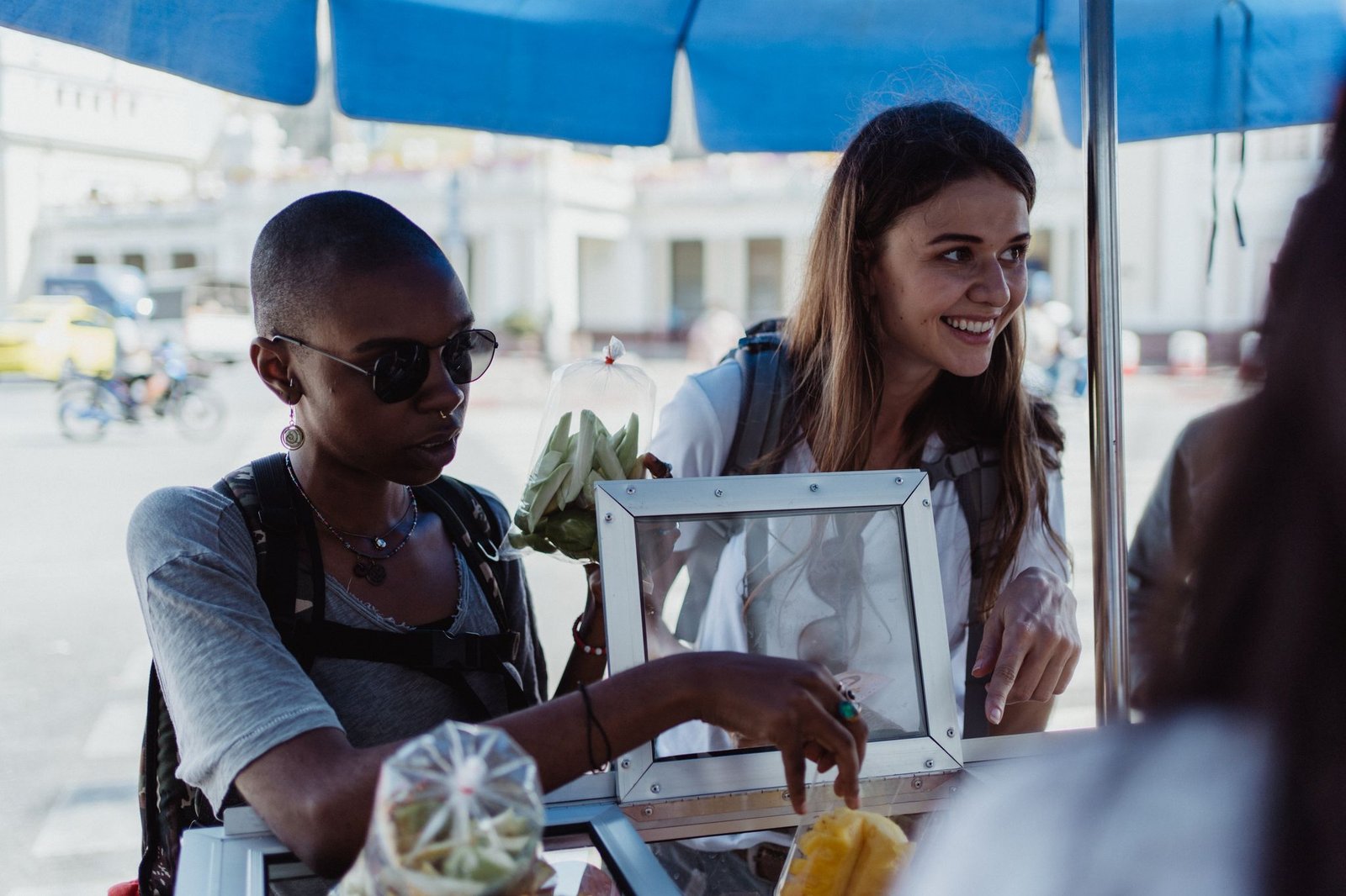 Two women happily purchasing snacks from a street food vendor under a blue canopy.