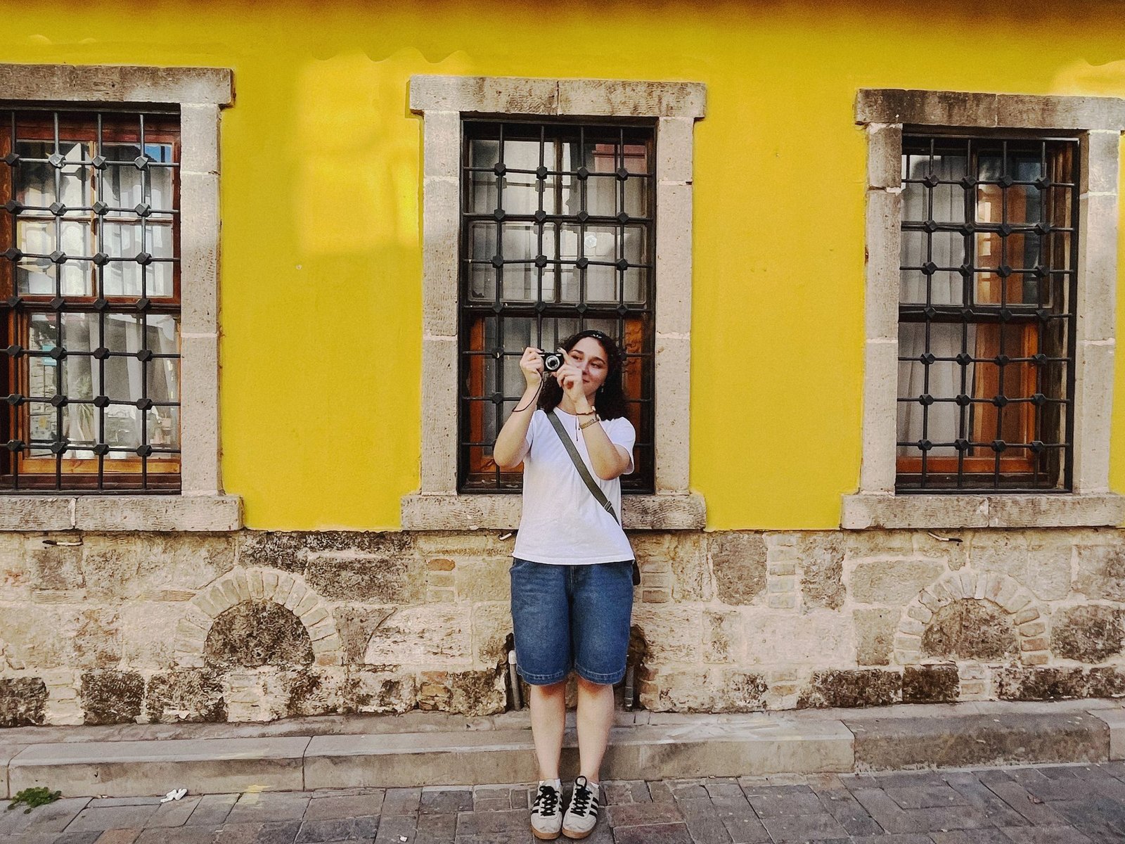 Female photographer taking a picture in front of a bright yellow wall with stone accents.